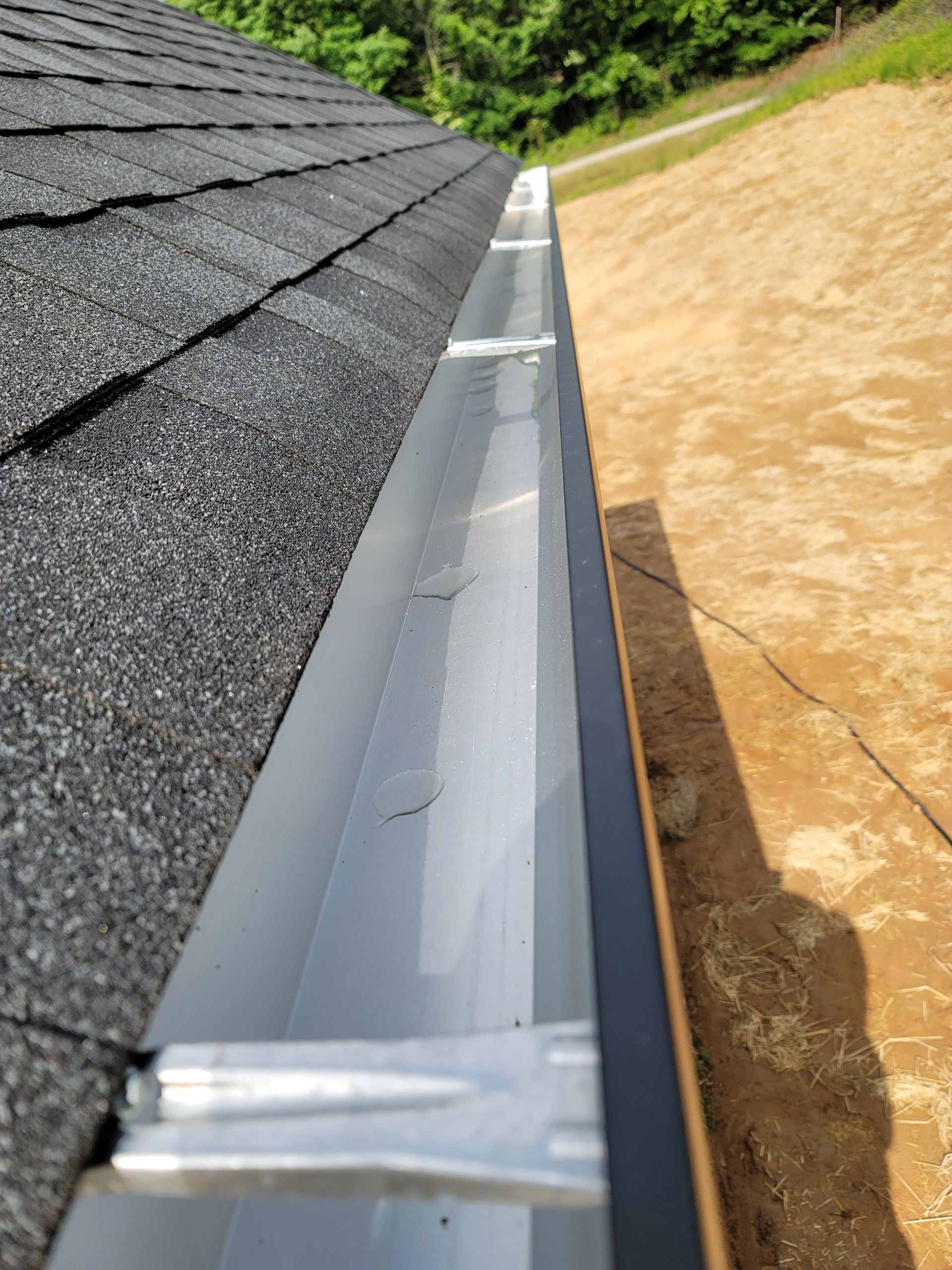 Close-up of roof and gutter. Gray shingles meet a metal gutter. Gutter's edge is black. Brown soil in the background.