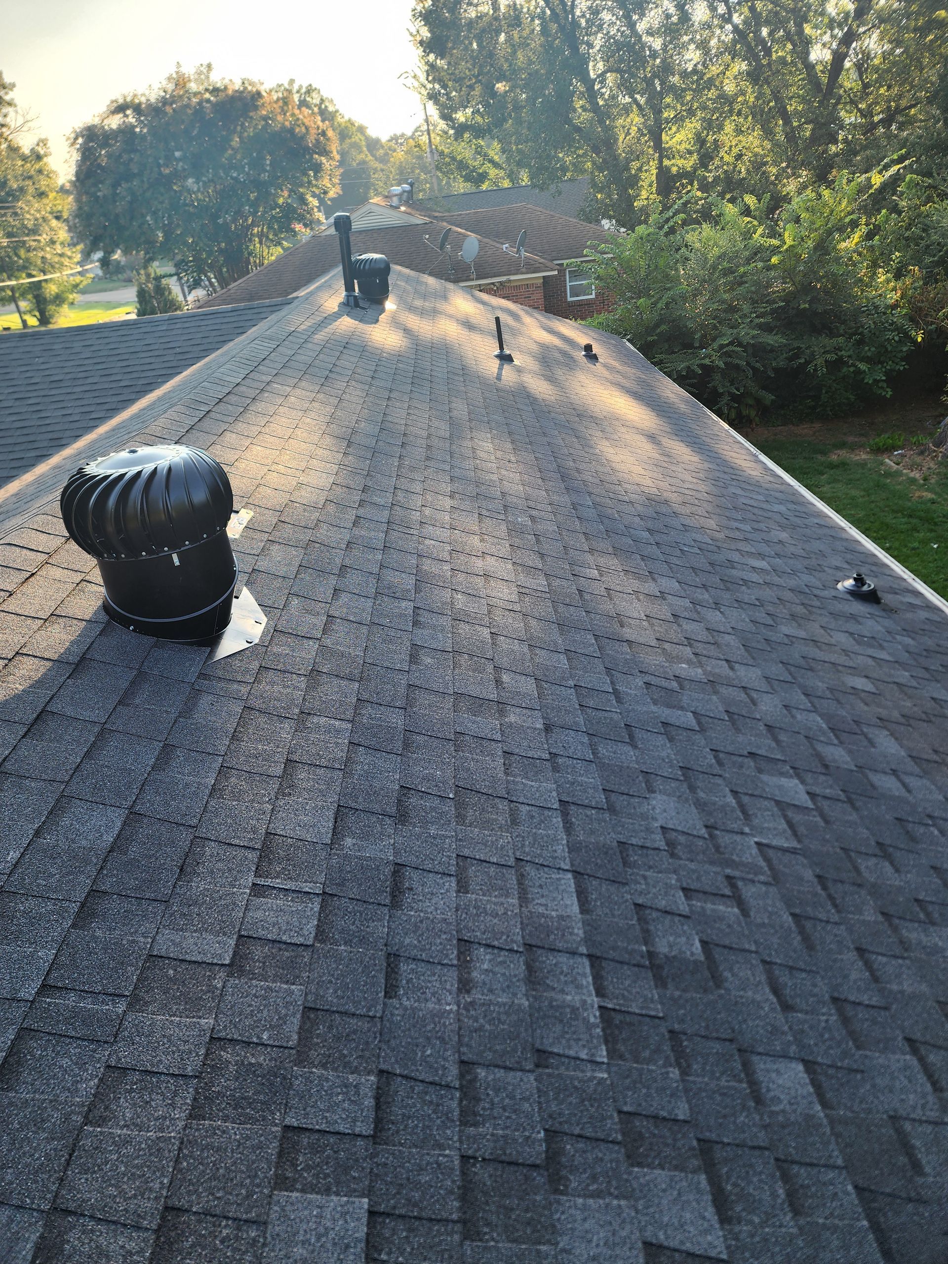 Dark gray shingled roof with a large vent, several smaller vents, and a few trees in the background.