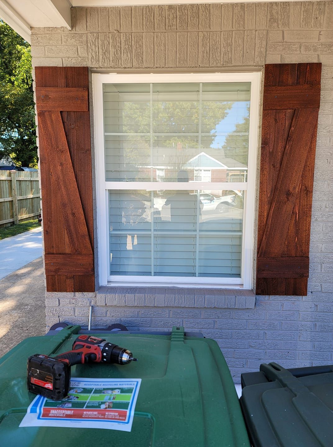 Brown wooden shutters flank a white-framed window on a brick wall. A drill rests on a green trash bin below.