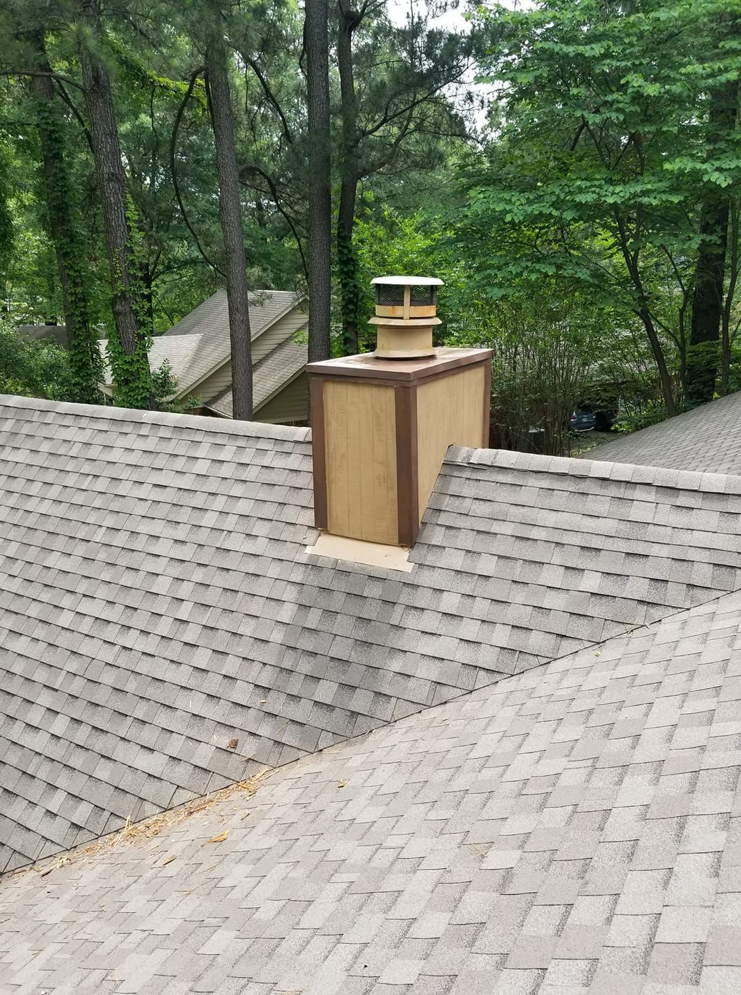 Chimney on a gray shingled roof, surrounded by trees. The chimney has a light brown finish and a cap.
