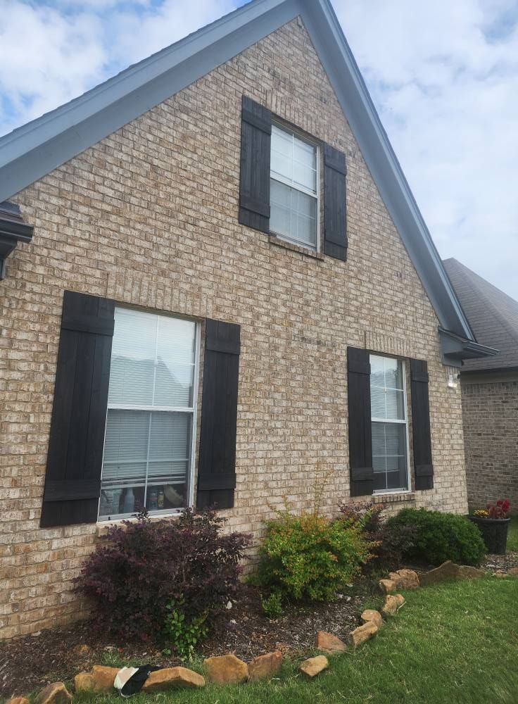 Tan brick house with black shutters, three windows visible, green landscaping.