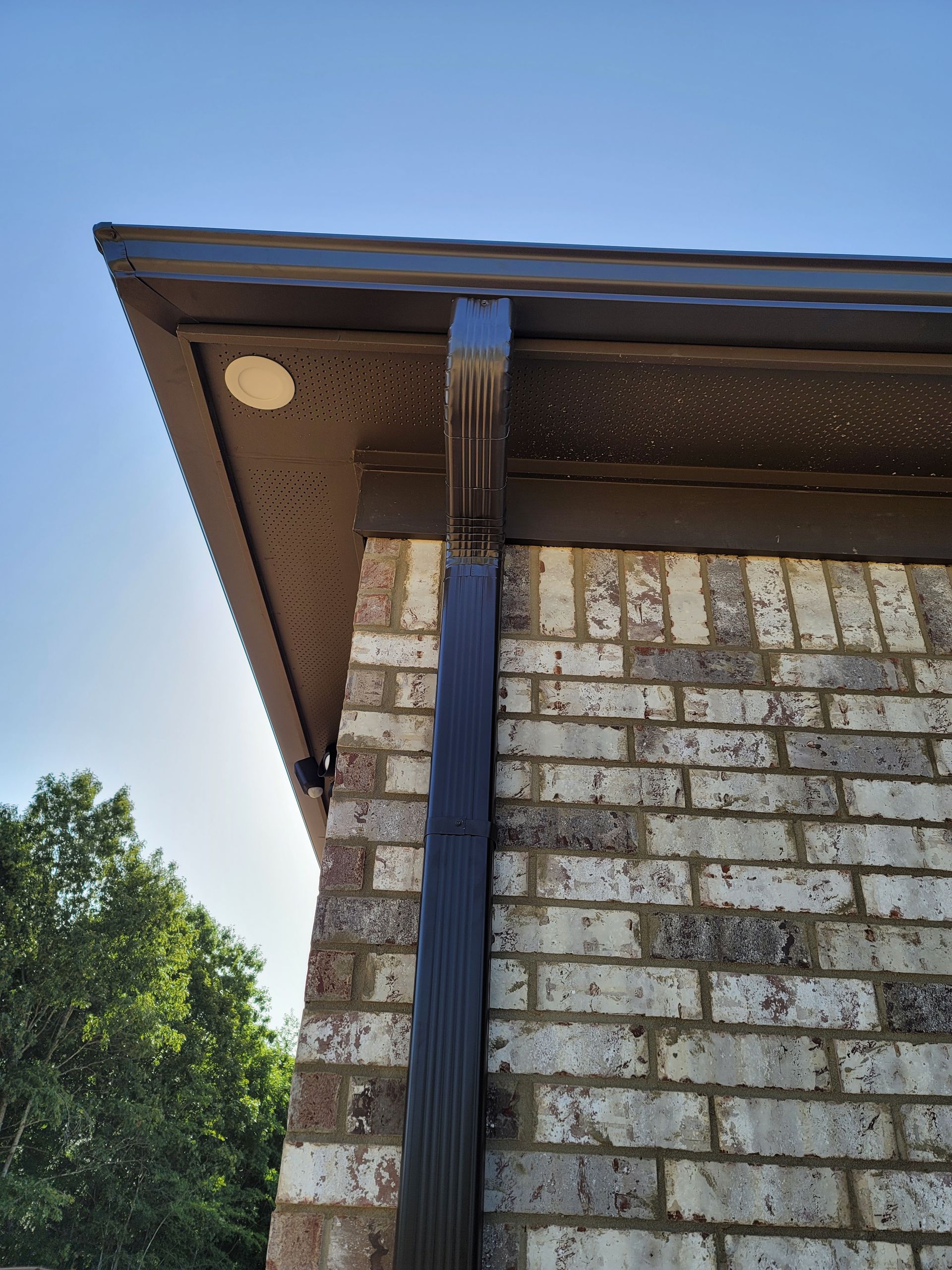 Black gutter downspout on a brick wall, with a dark brown eave and a blue sky backdrop.