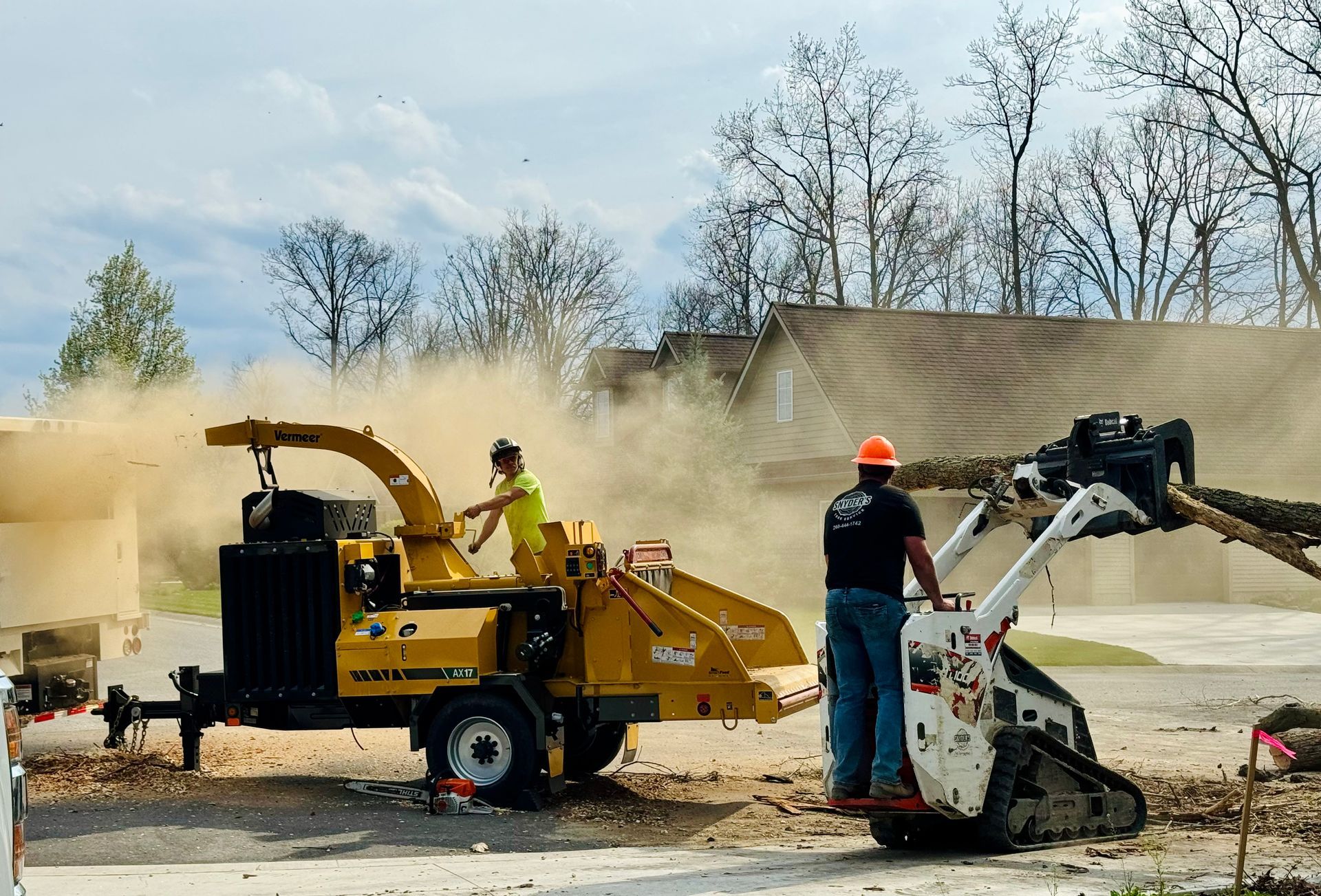 Two workers using a wood chipper to process tree branches. One loads the machine; the other operates a skid steer.