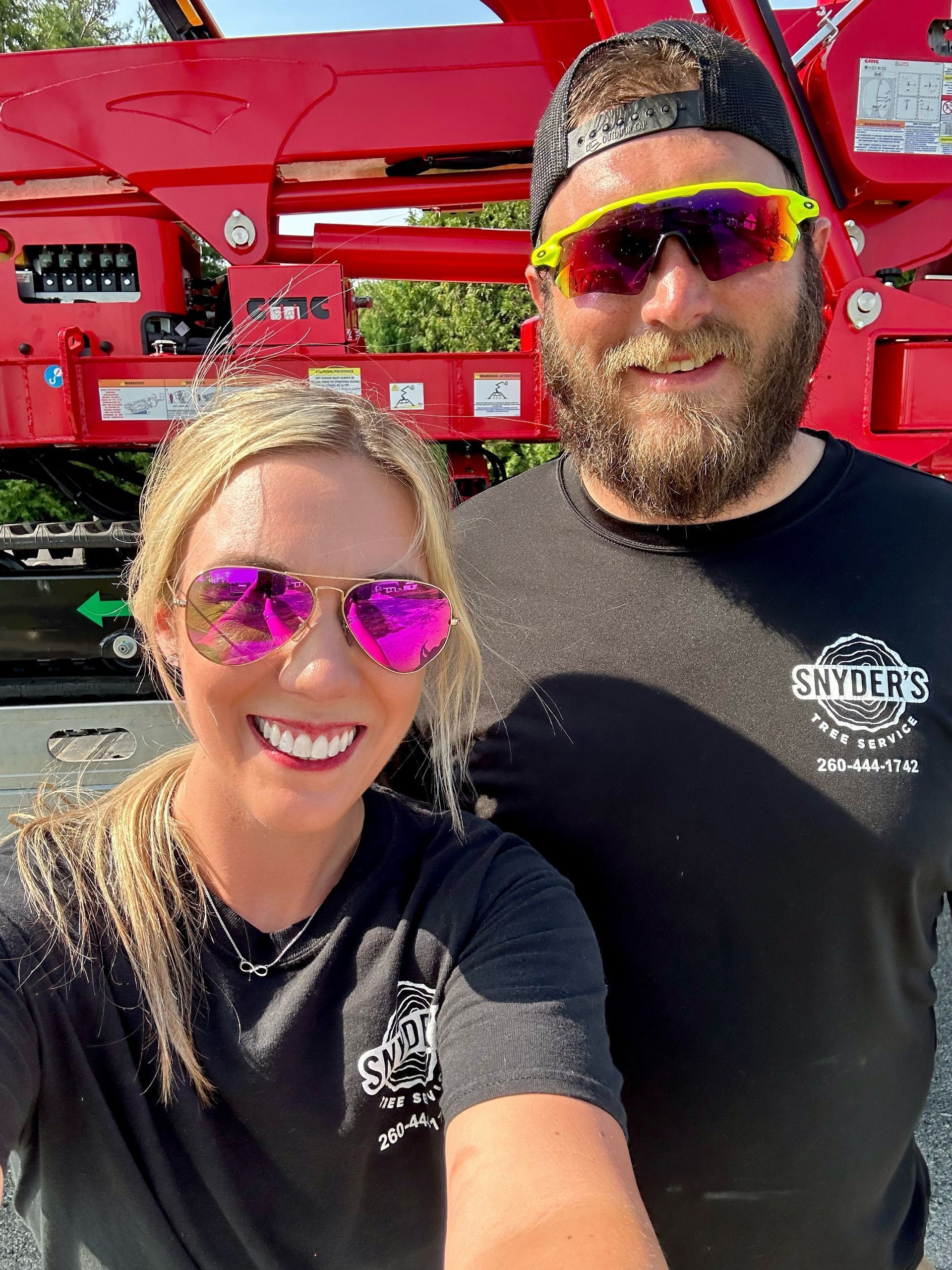 Two people wearing sunglasses smile in front of red machinery. Both wear black shirts with the same logo.