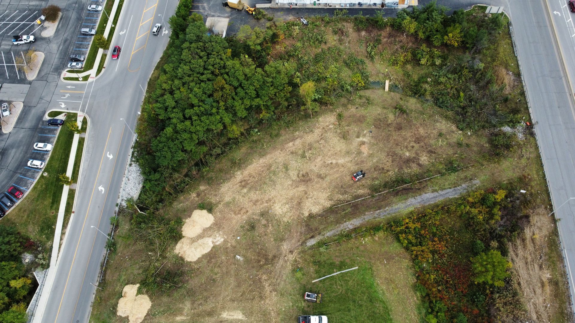 Aerial view of an empty lot with patches of trees, grass, and a paved road on either side.