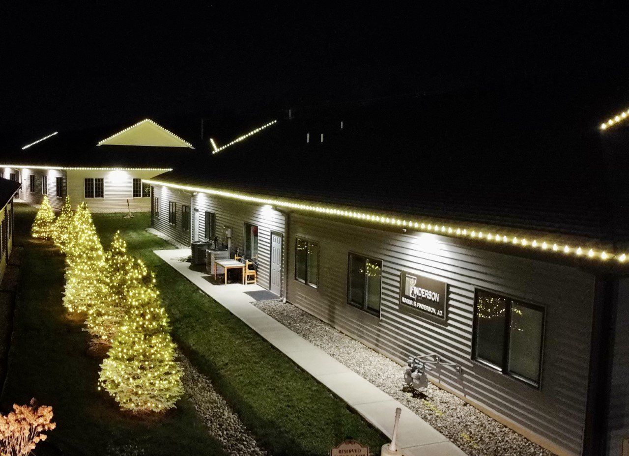 Building exterior at night, decorated with Christmas lights on the roof and trees, with a lit walkway.