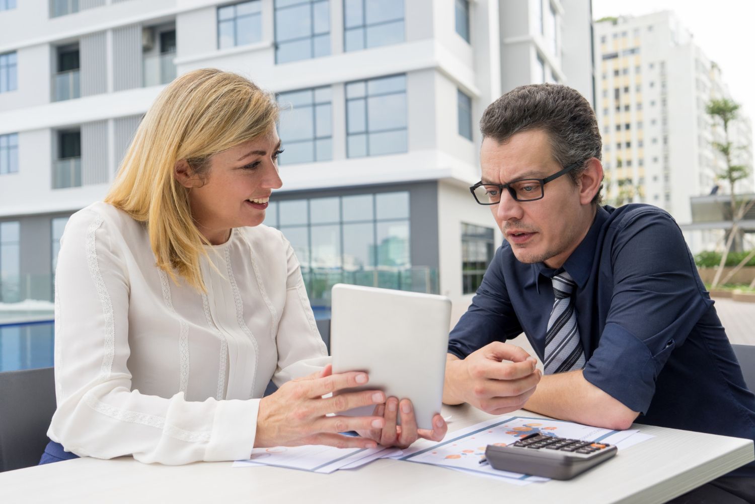 Two people reviewing documents at an outdoor table with a calculator and tablet in front of a modern office building