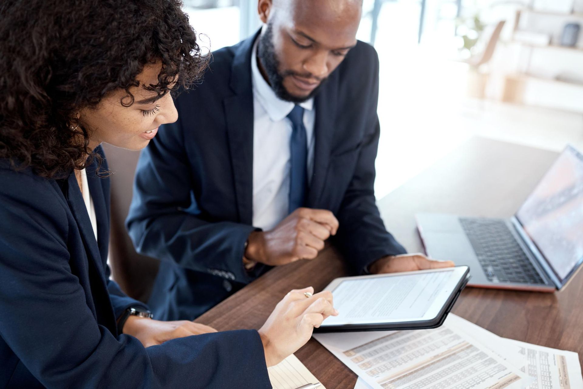 Two coworkers in suits review a tablet and papers beside a laptop at a desk