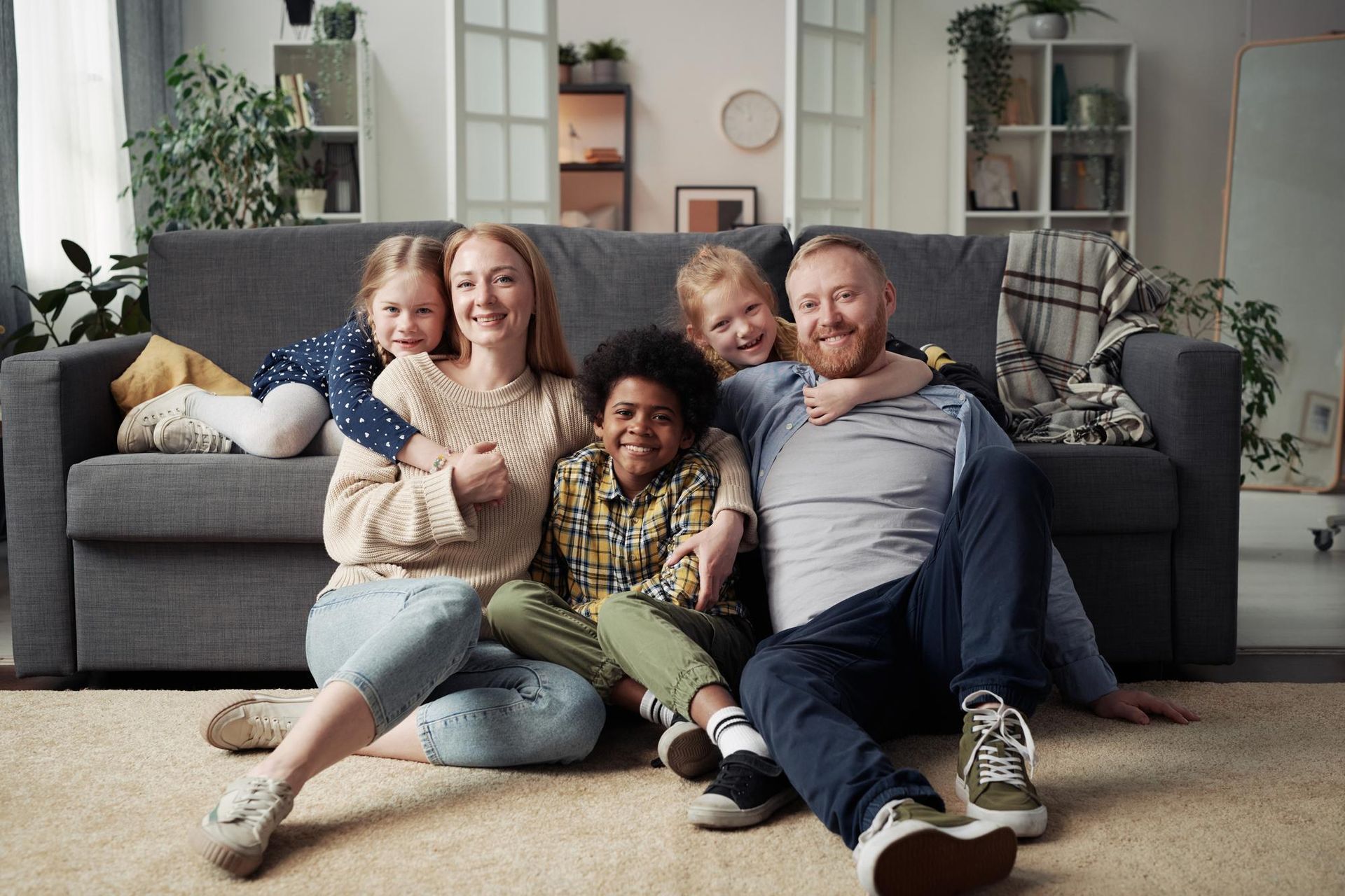 Four people sitting on the floor in a living room, smiling in front of a gray couch.