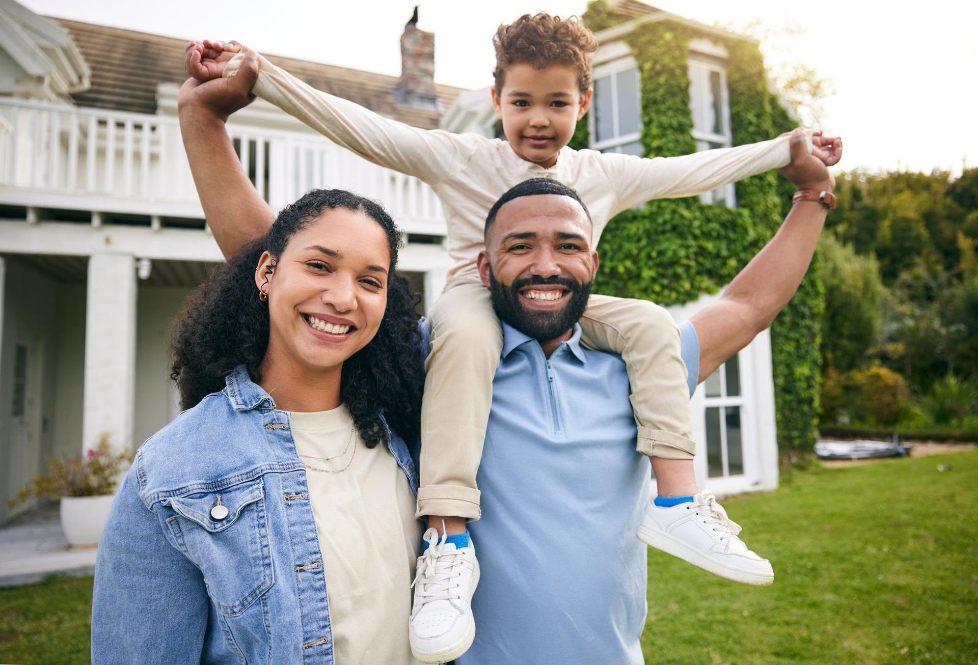 Family smiling outdoors, child on father’s shoulders, in front of a house with a green lawn.