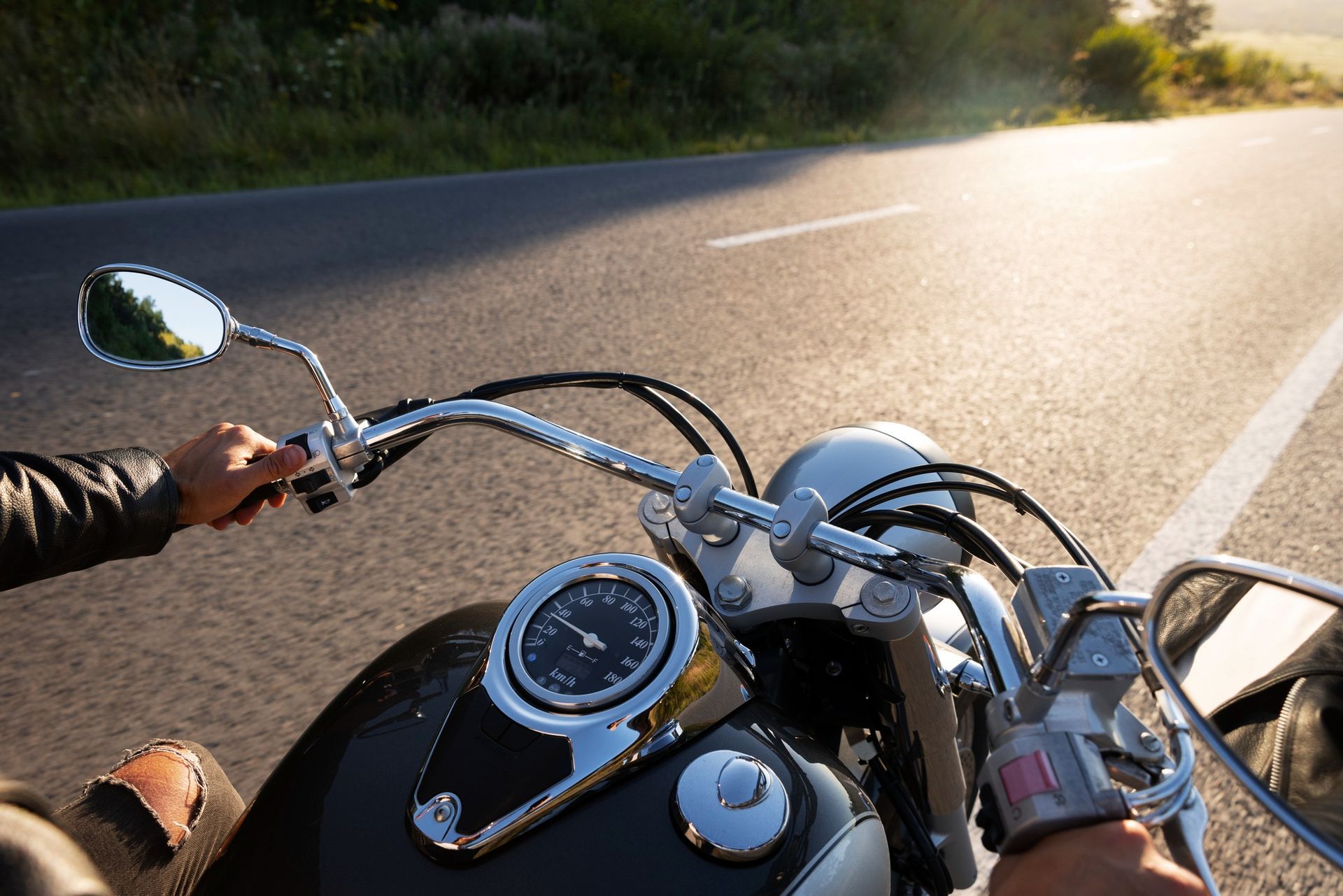 Motorcycle handlebars and dashboard on a sunlit road, with a rider’s hands visible