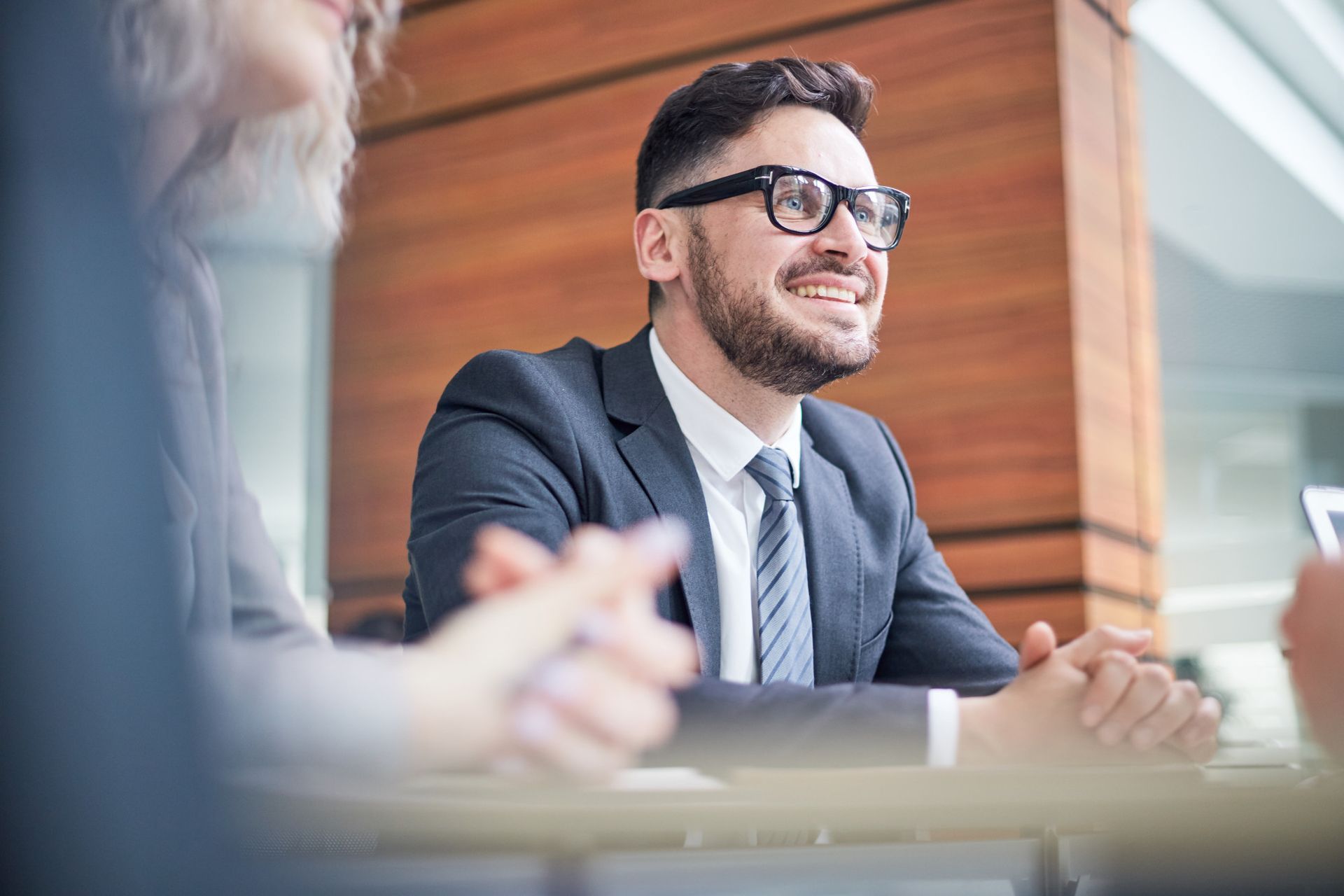 Businessman smiling in a meeting room, wearing glasses and a suit, with colleagues blurred in the foreground