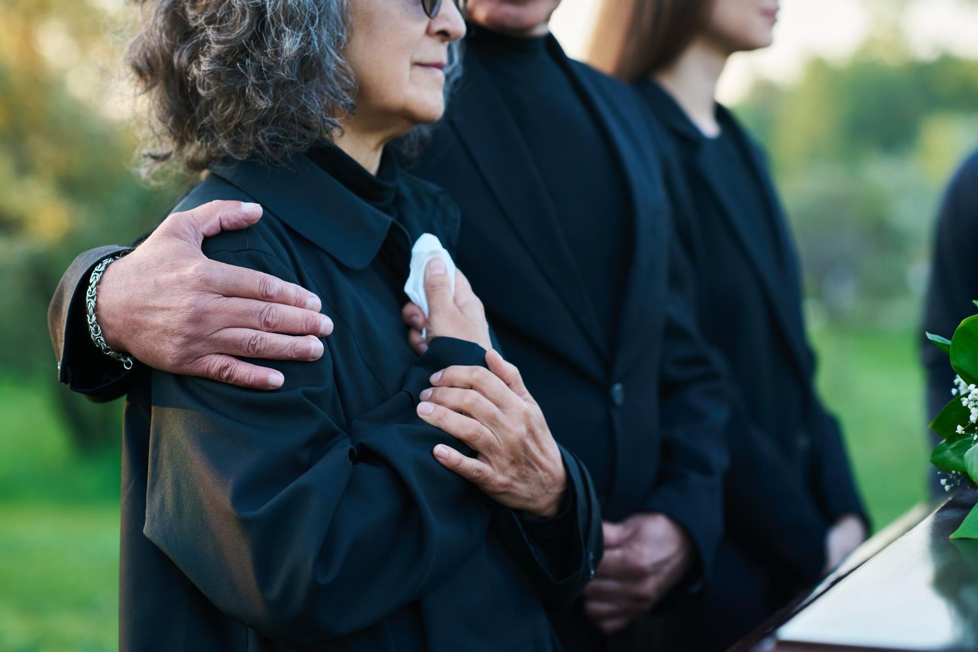 Mourners in black embrace at an outdoor memorial beside flowers and a casket