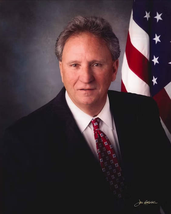 Formal portrait of a man in a suit and red tie beside a U.S. flag
