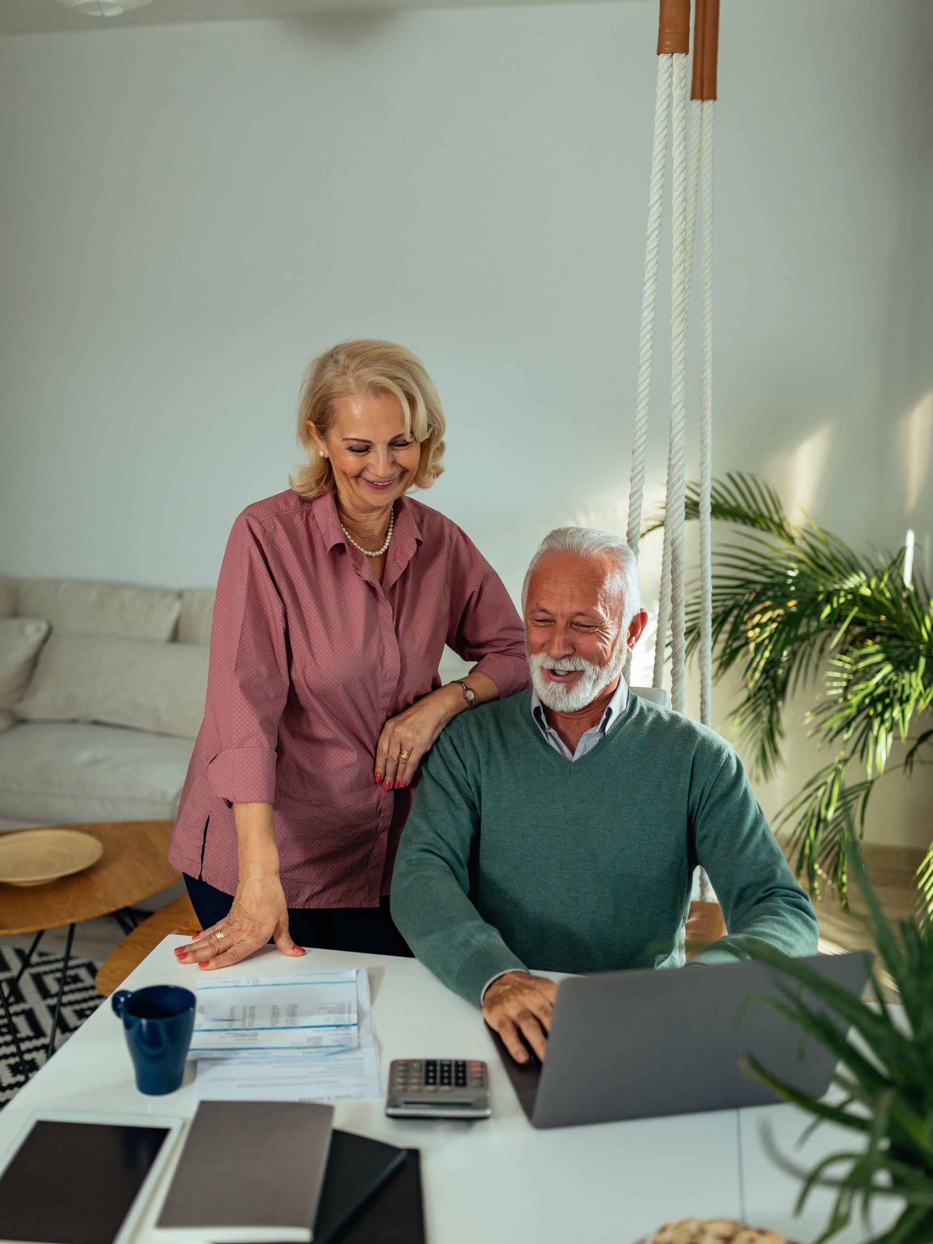 Two people smiling at a home office desk with a laptop, papers, and a cup in a bright room