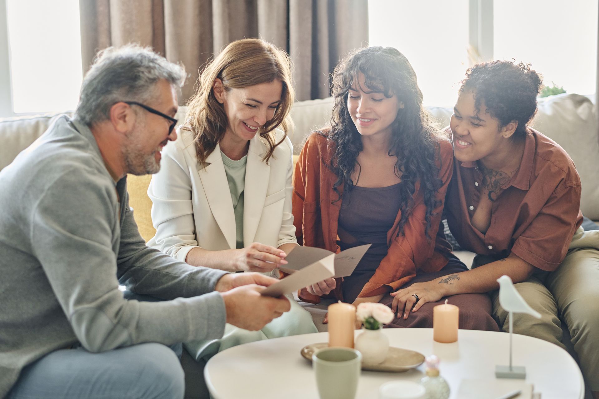 Four people sit on a couch and chair, smiling and reading a card together in a cozy living room.