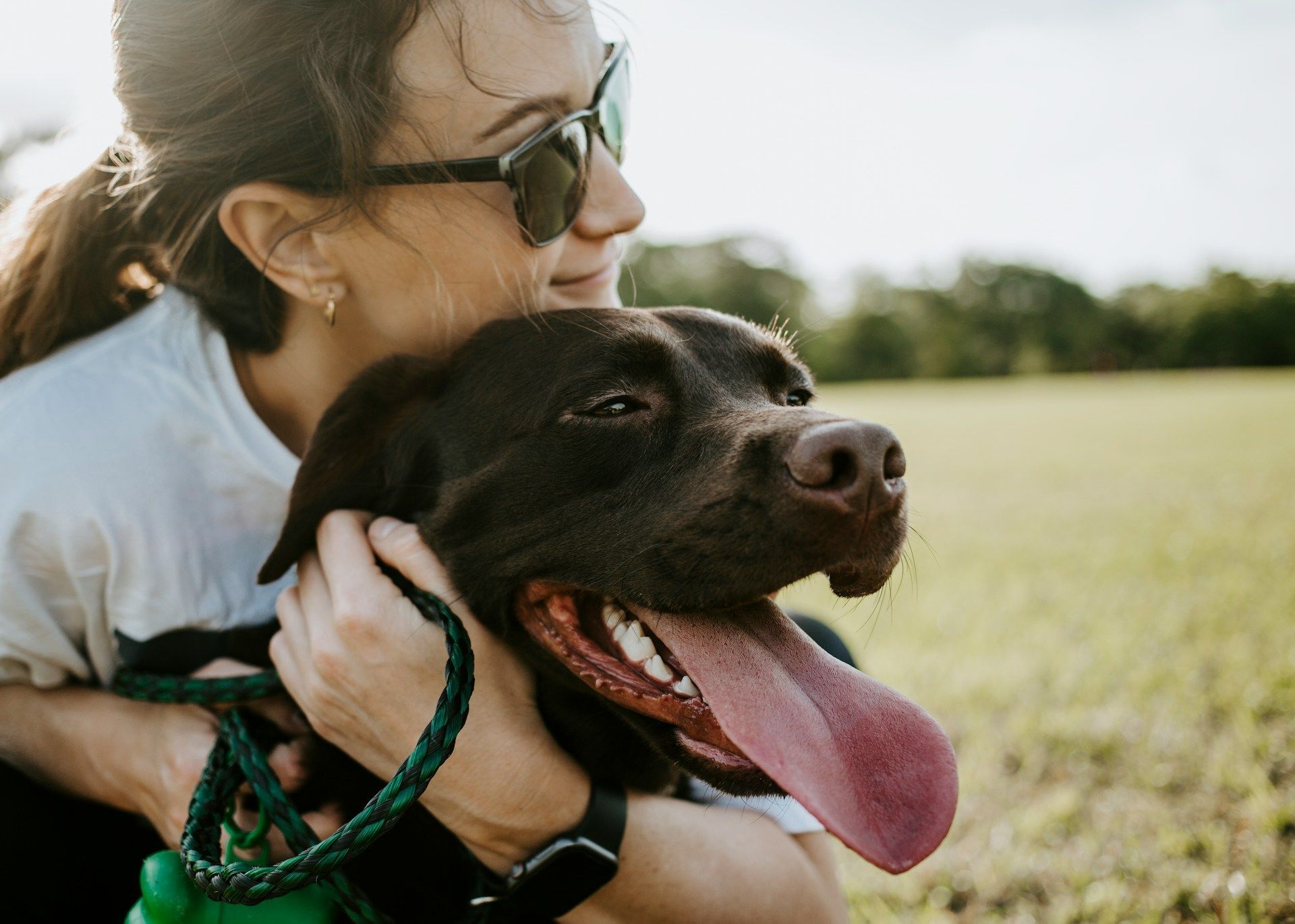 Woman hugging a black dog with a green leash in a sunny field, both with tongues out