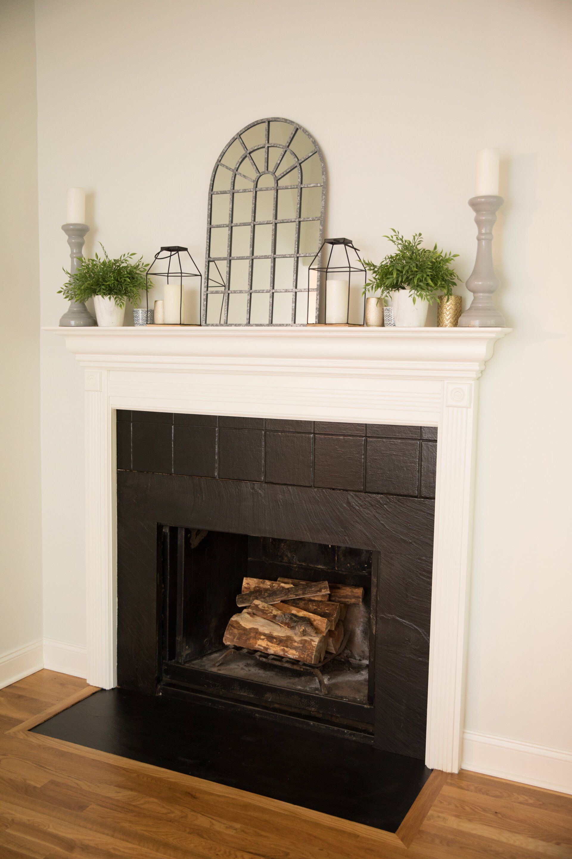 Fireplace with black brick, white mantel with decor: mirror, lanterns, plants, candles.