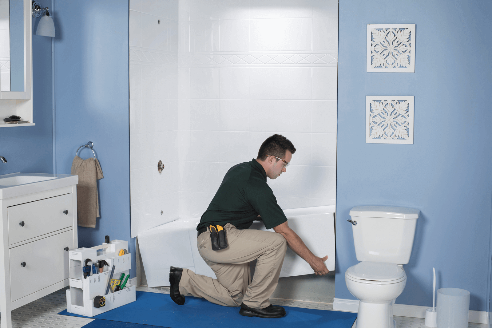 Man installing a white bathtub surround in a blue bathroom; tools and a toilet are visible.