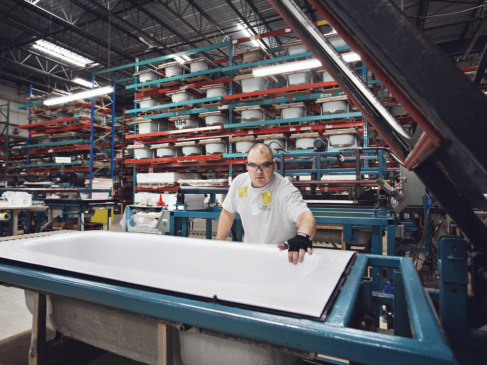 Man working on a large, white bathtub in a factory. Shelves of bathtubs are in the background.