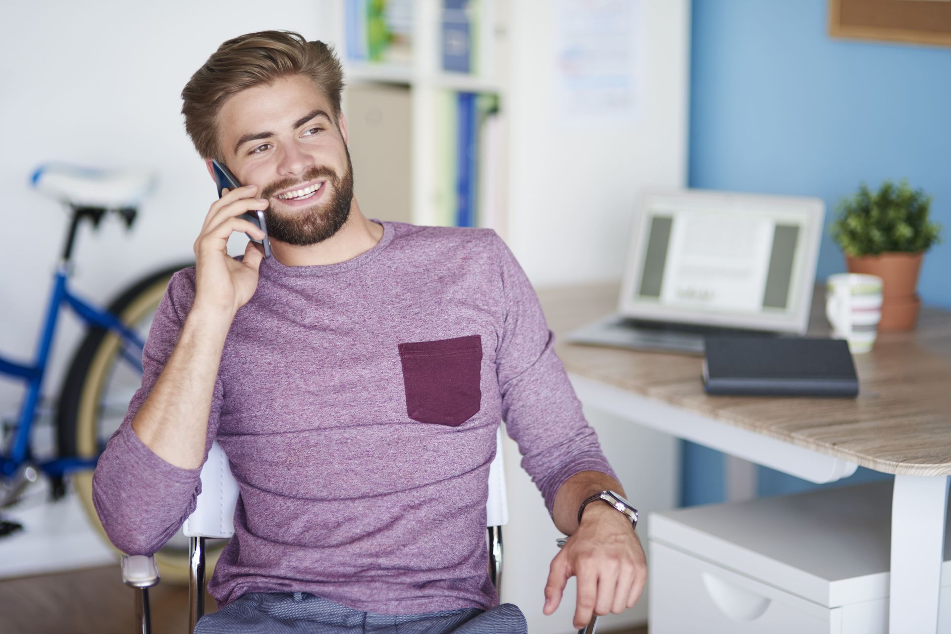 Man with beard, talking on a cell phone, smiling, sitting at a desk with a laptop and a bicycle in the background.