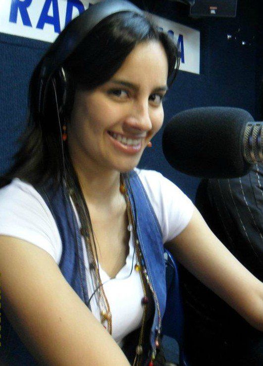 Mujer sonriendo, con auriculares, sentada frente a un micrófono en un estudio de radio.