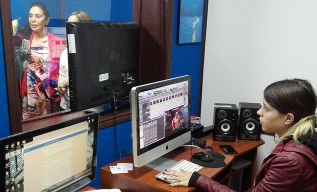 Mujer editando video en la computadora, con monitores en el escritorio. Otra mujer se refleja en la ventana detrás de ella.