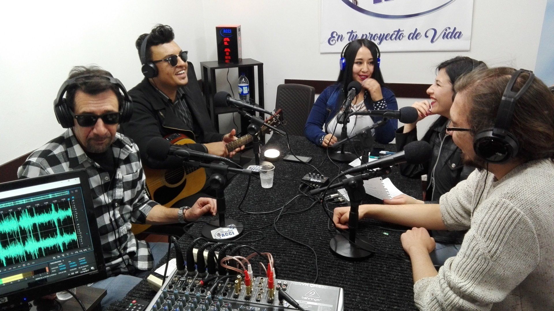Personas en un estudio de grabación. Un hombre toca la guitarra. Otros están sentados frente a los micrófonos, sonriendo. Se ve el equipo de mezcla de sonido.