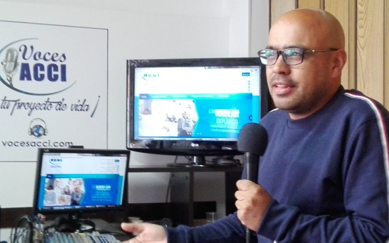 Hombre hablando por micrófono, frente a monitores que muestran un sitio web y el logo 