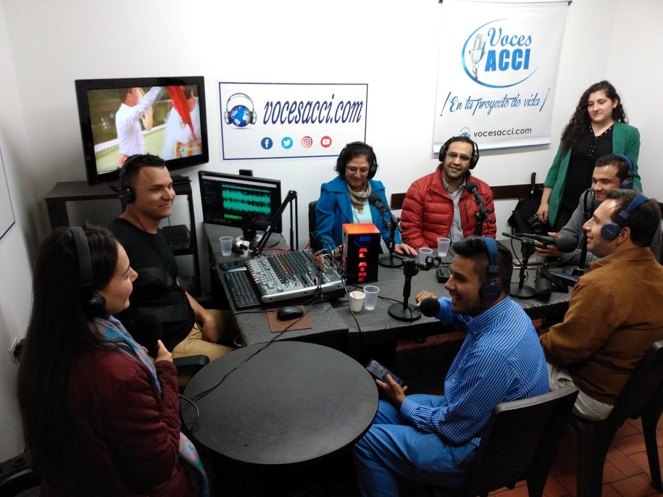 Grupo de personas en una estación de radio, sentados alrededor de micrófonos y una mesa de sonido.