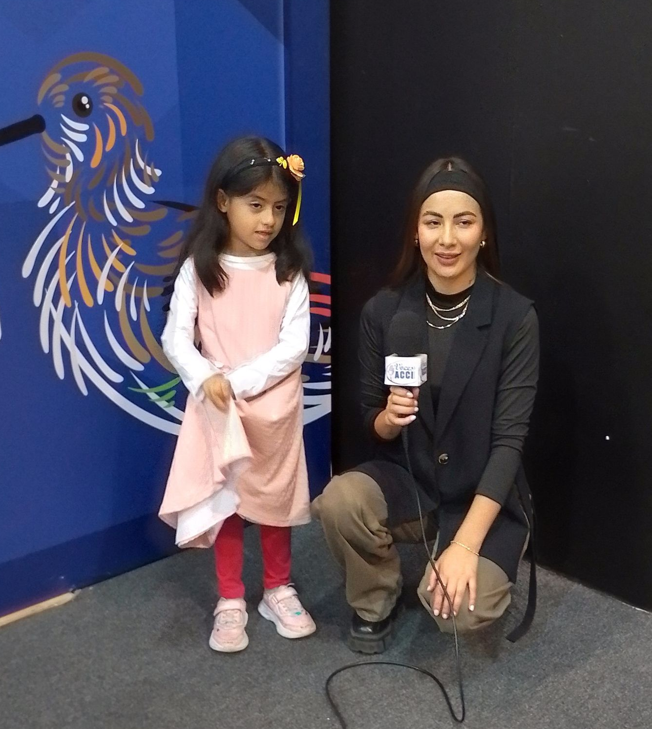 Un niño y una mujer con un micrófono se encuentran frente a una pared azul con un diseño de colibrí.