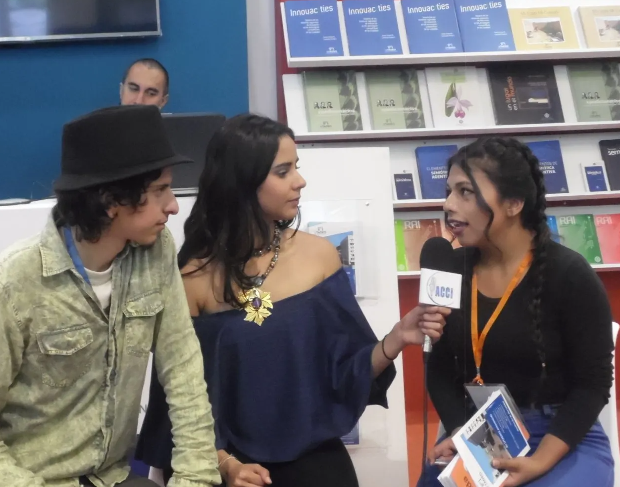 Una mujer entrevista a dos personas en una feria del libro. El entrevistador sostiene un micrófono y los entrevistados la miran.