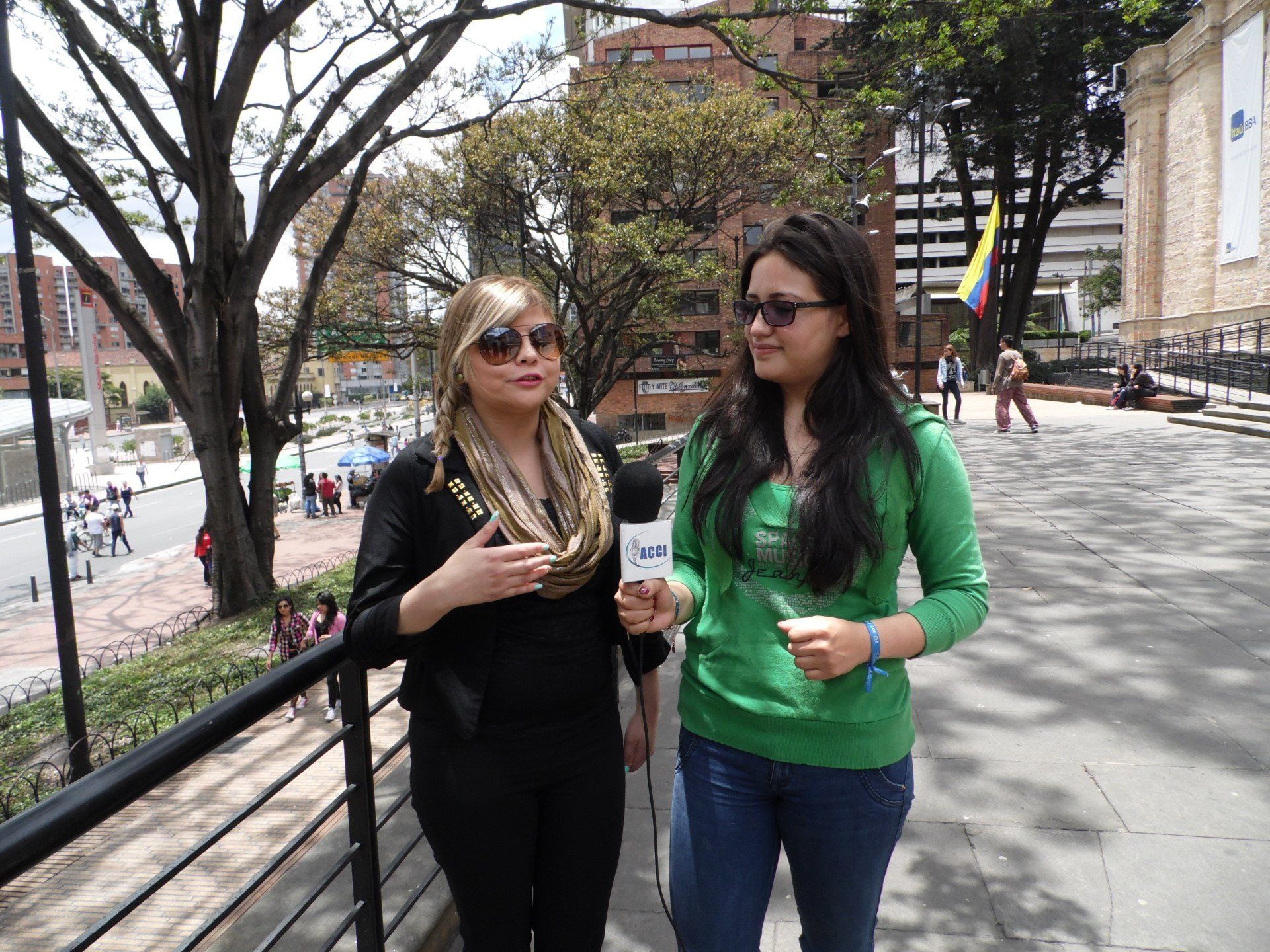 Dos mujeres afuera, una con un micrófono. Edificios y árboles al fondo.