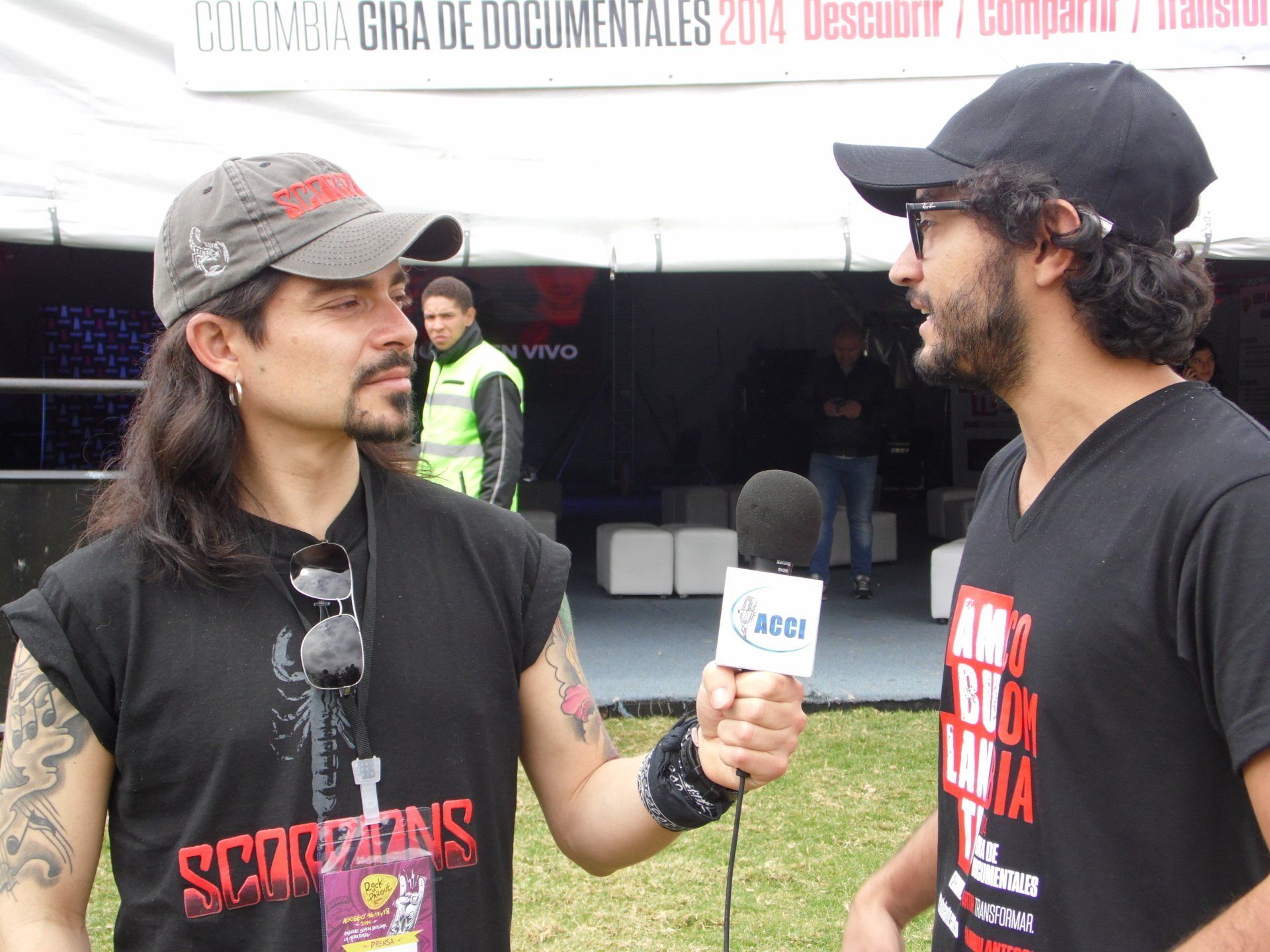 Dos hombres conversando, uno con un micrófono. Al aire libre, bajo una carpa. Uno lleva una camiseta de los Scorpions.