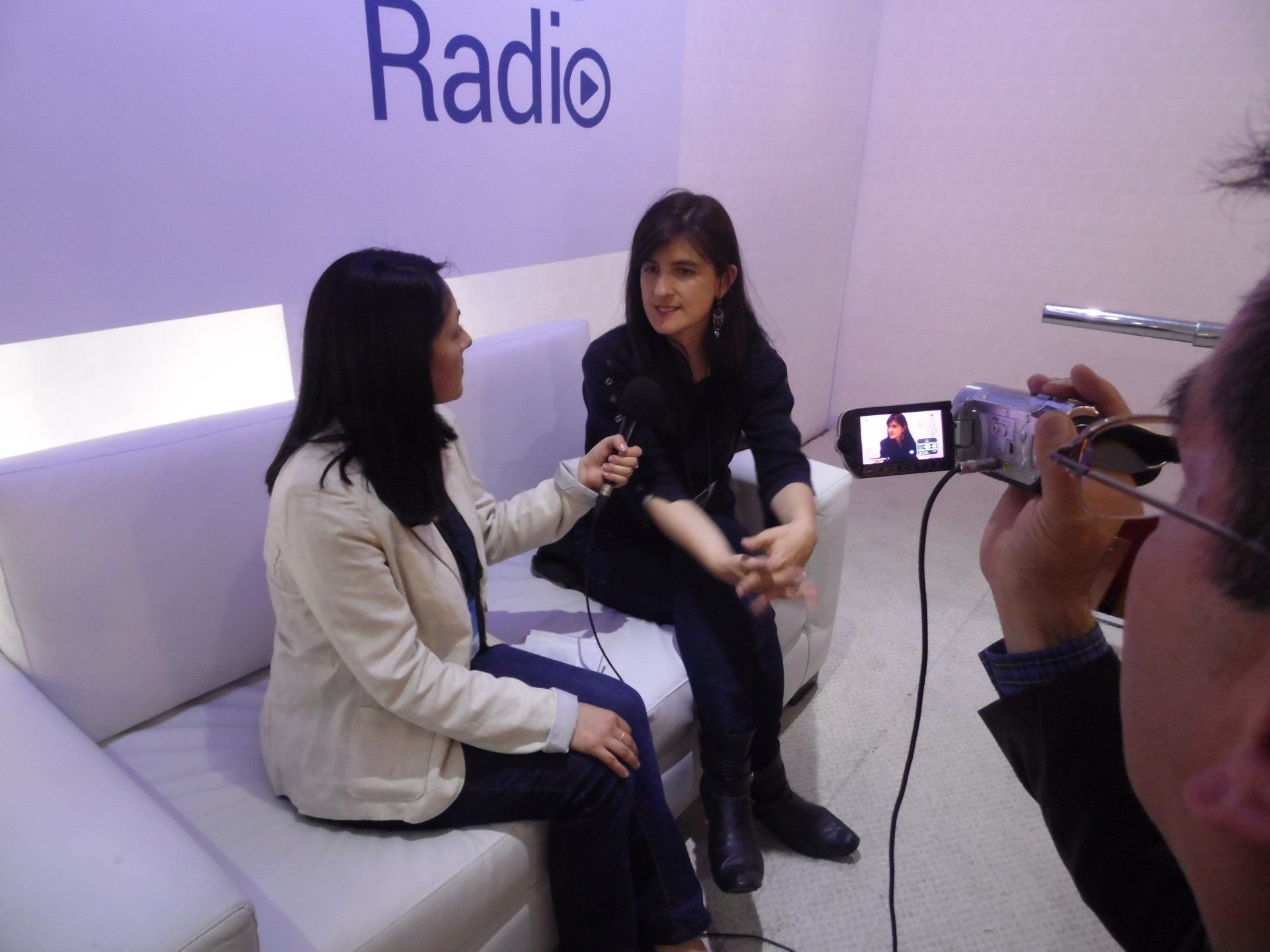 Mujer entrevistando a otra mujer en un sofá blanco; una cámara está grabando la entrevista.