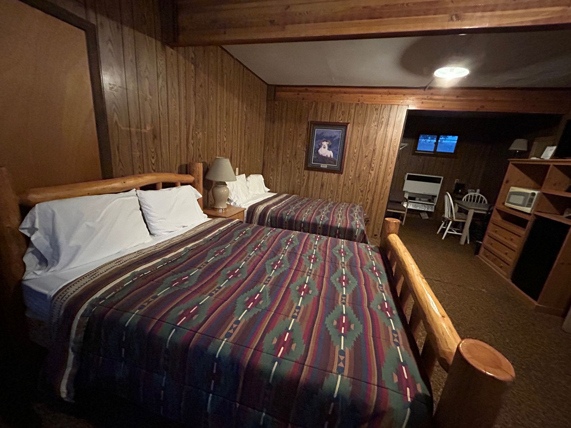 Two beds in a rustic room with wood paneling. Additional living area visible beyond.