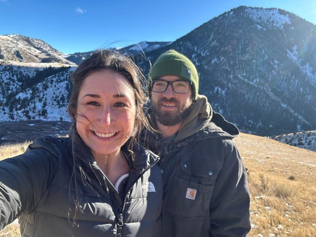 Woman and man smiling for a selfie in a mountain landscape, blue sky, dry grass, man wearing a green beanie.