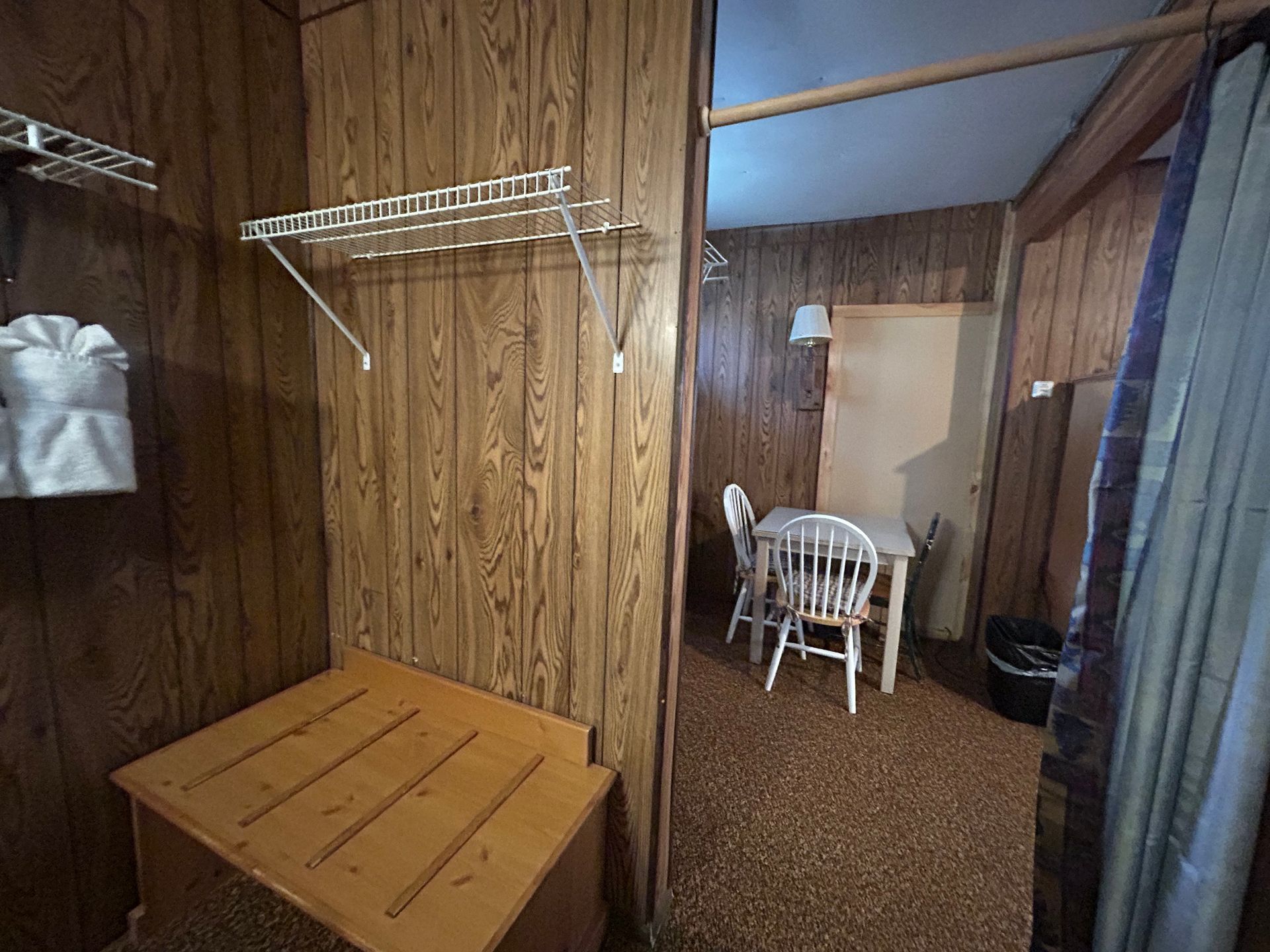 Wooden-paneled motel room. Bench, shelf with towels, table, and chairs visible through doorway.