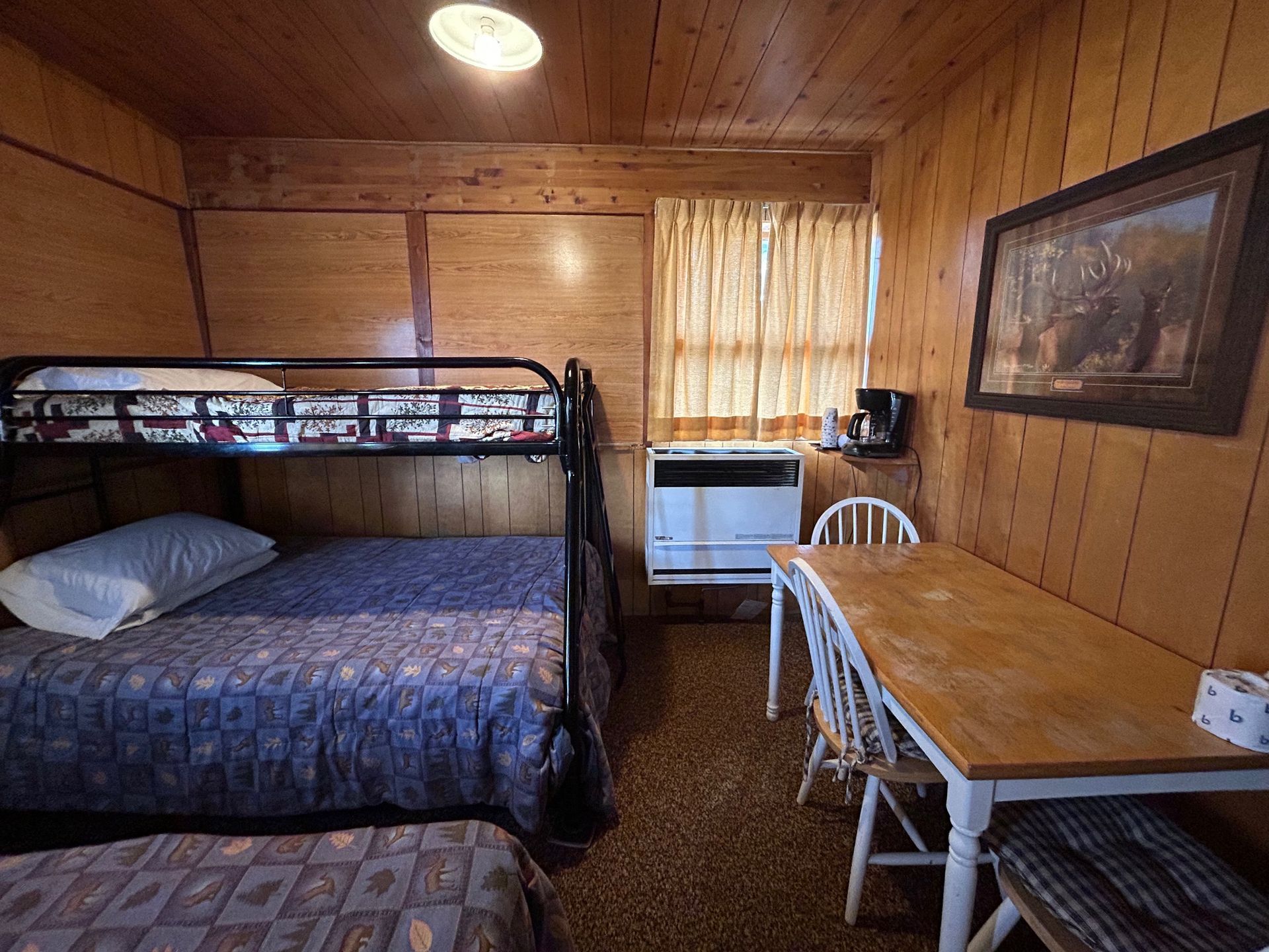 Interior of a rustic cabin with bunk beds, a table, a window, and wood-paneled walls.