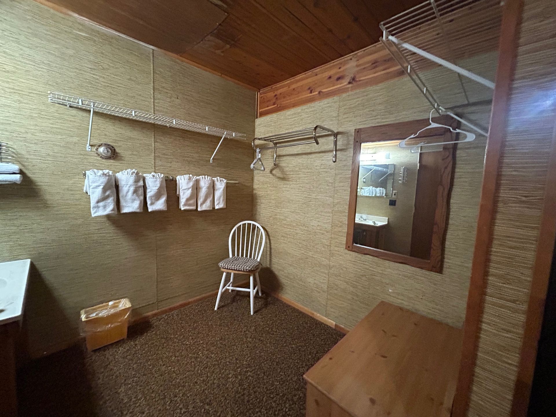 Small, rustic room with a chair, mirror, and clothes rack. Beige walls, wood trim, and brown carpet.