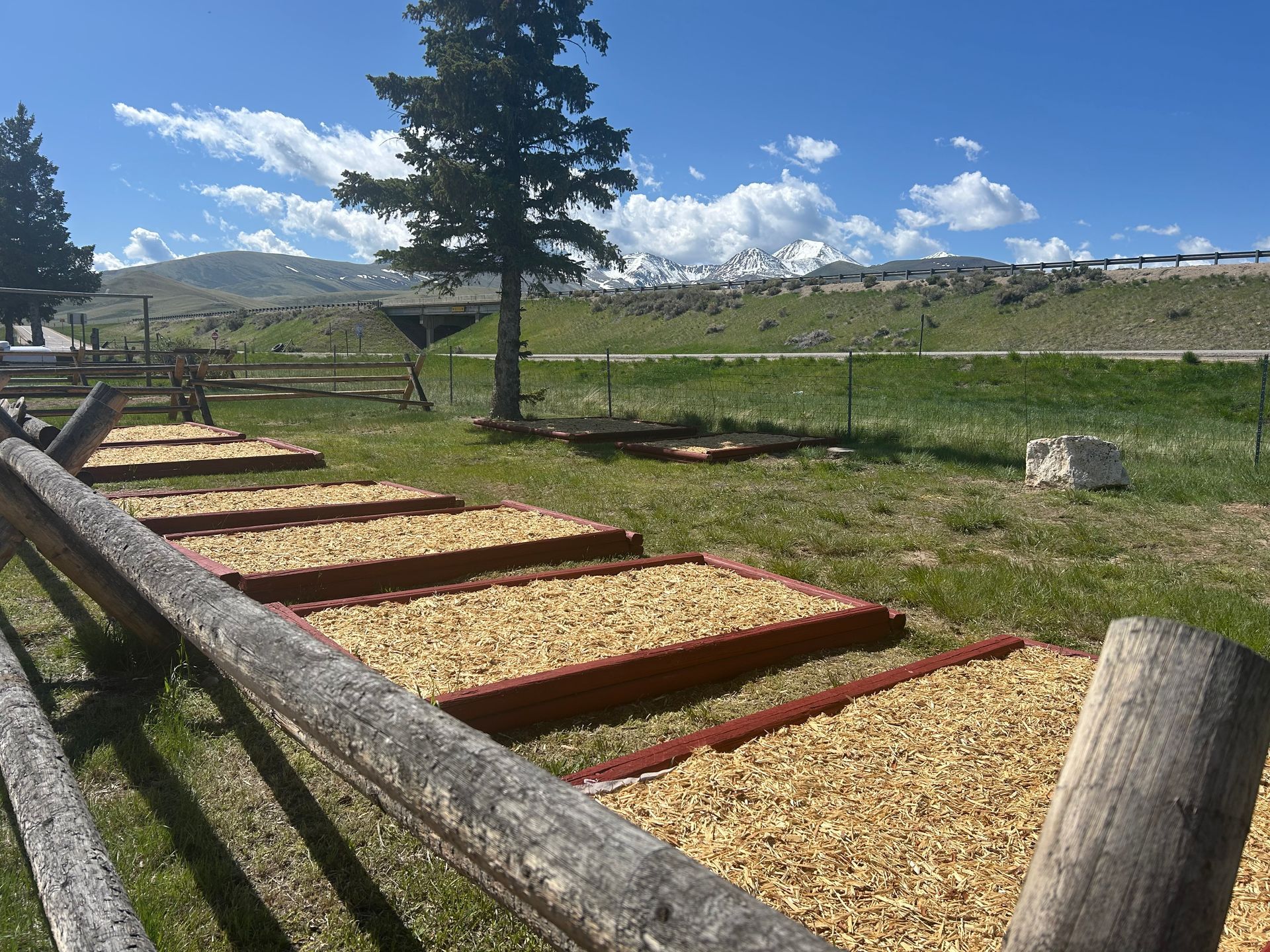 Wooden raised garden beds filled with wood chips, next to a weathered fence. Mountain landscape background.