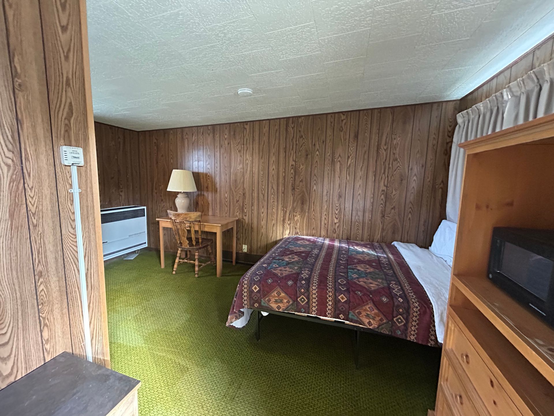 A hotel room with a bed, desk, and microwave. Wooden panel walls, green carpet.