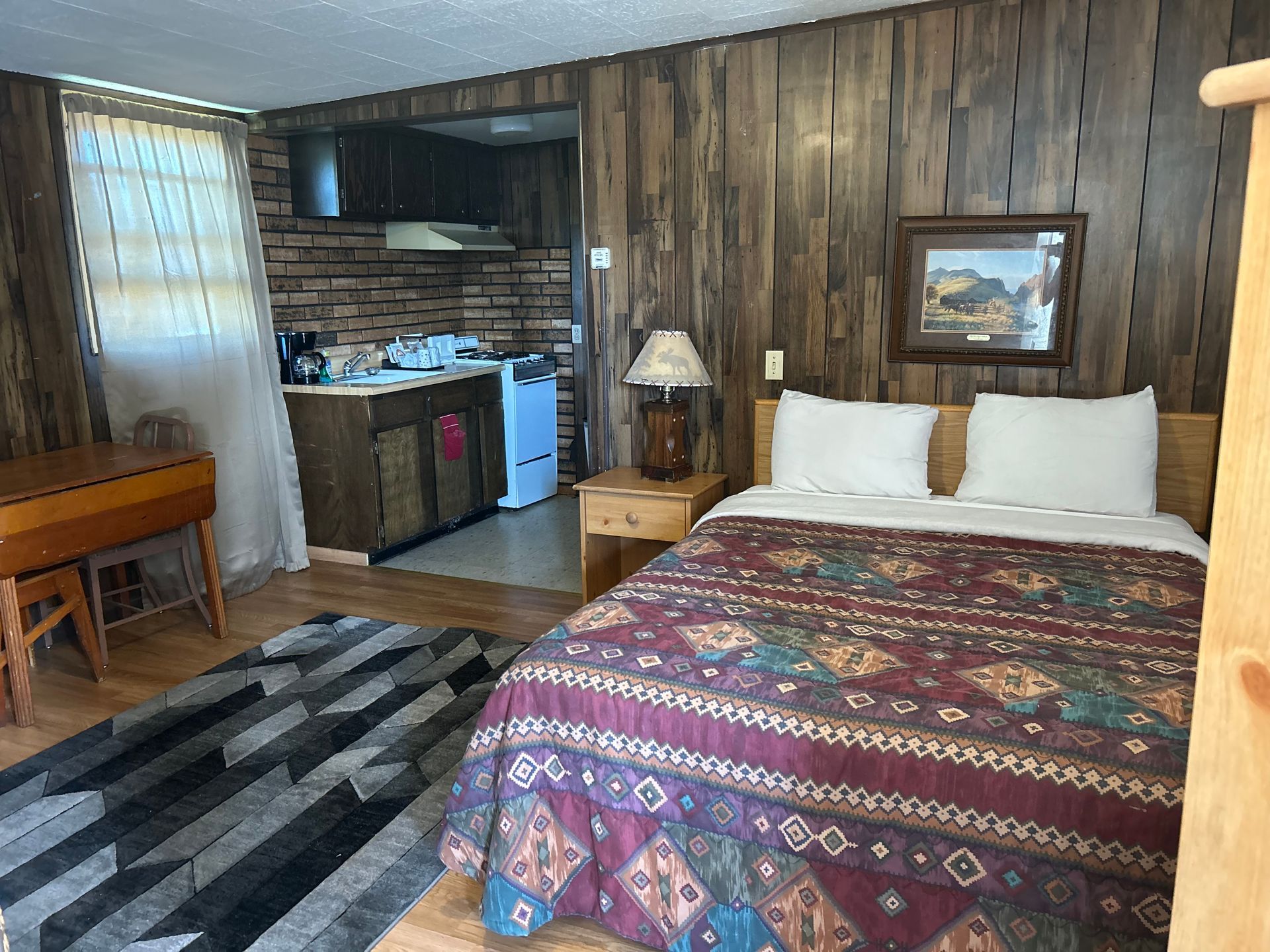 Bedroom with wooden panel walls, bed with patterned quilt, kitchenette visible in the background.