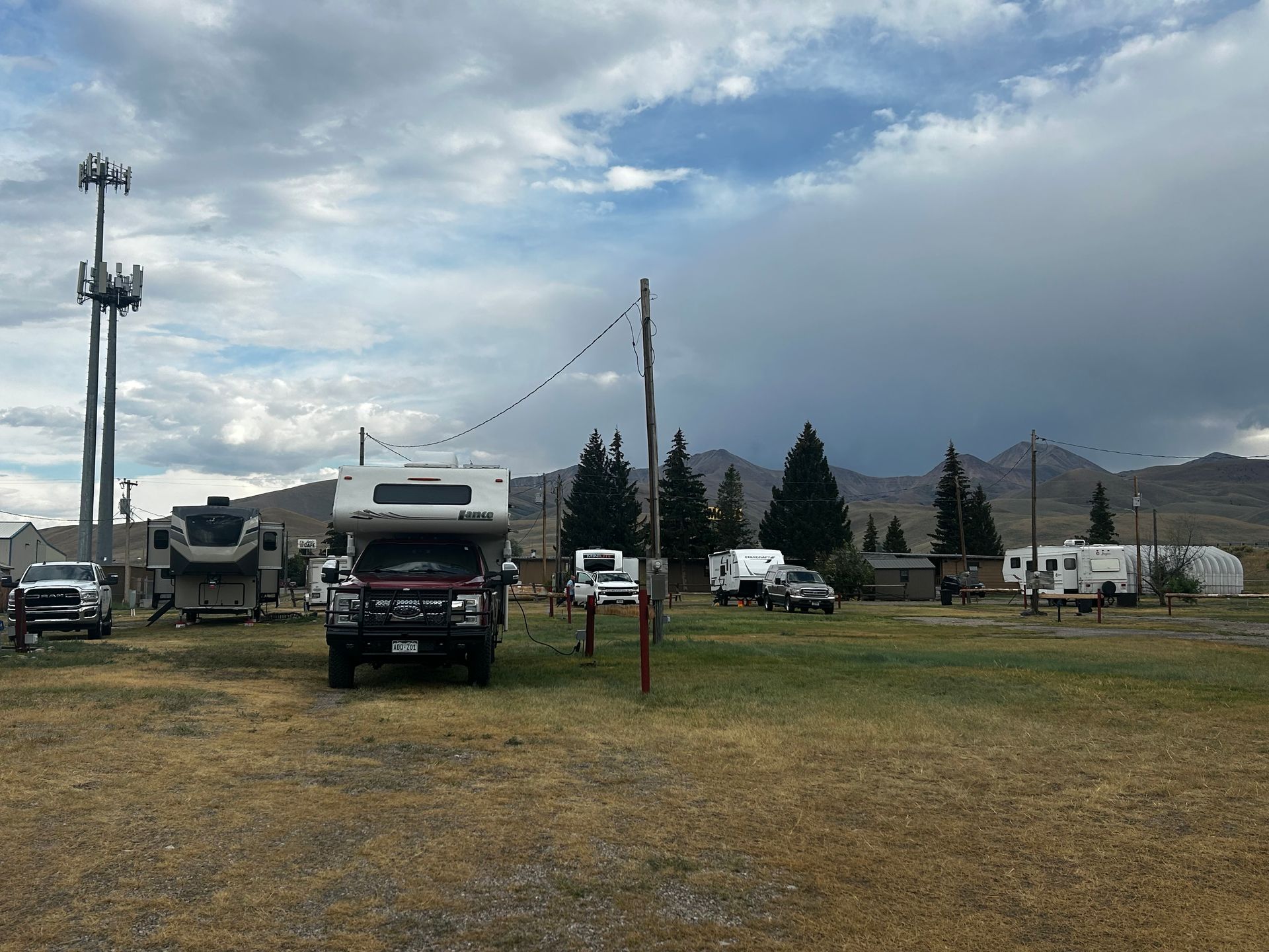 Campground with RVs, cell tower, string lights, and mountains under a cloudy sky.