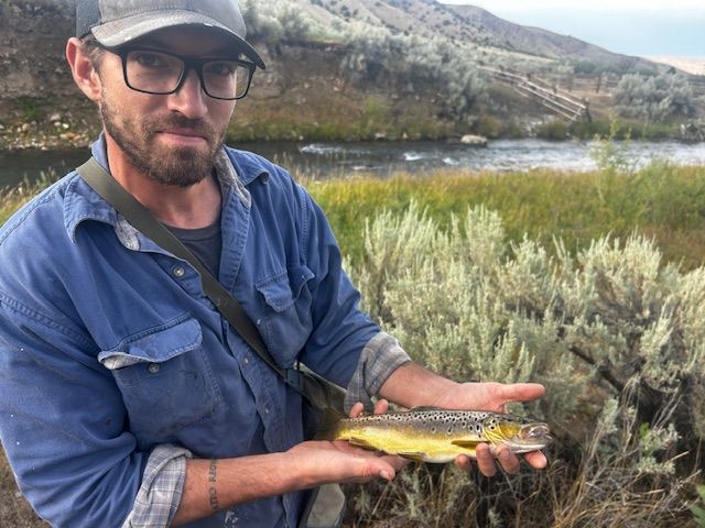 Man holding a brown trout by a river. He wears a cap and glasses, with a neutral expression.