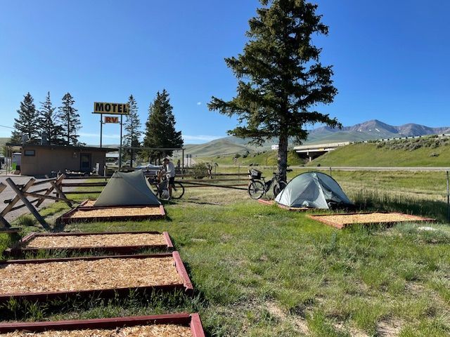 Two tents pitched at a motel campsite; a person stands near one bike. Blue sky, highway, and mountain in the distance.
