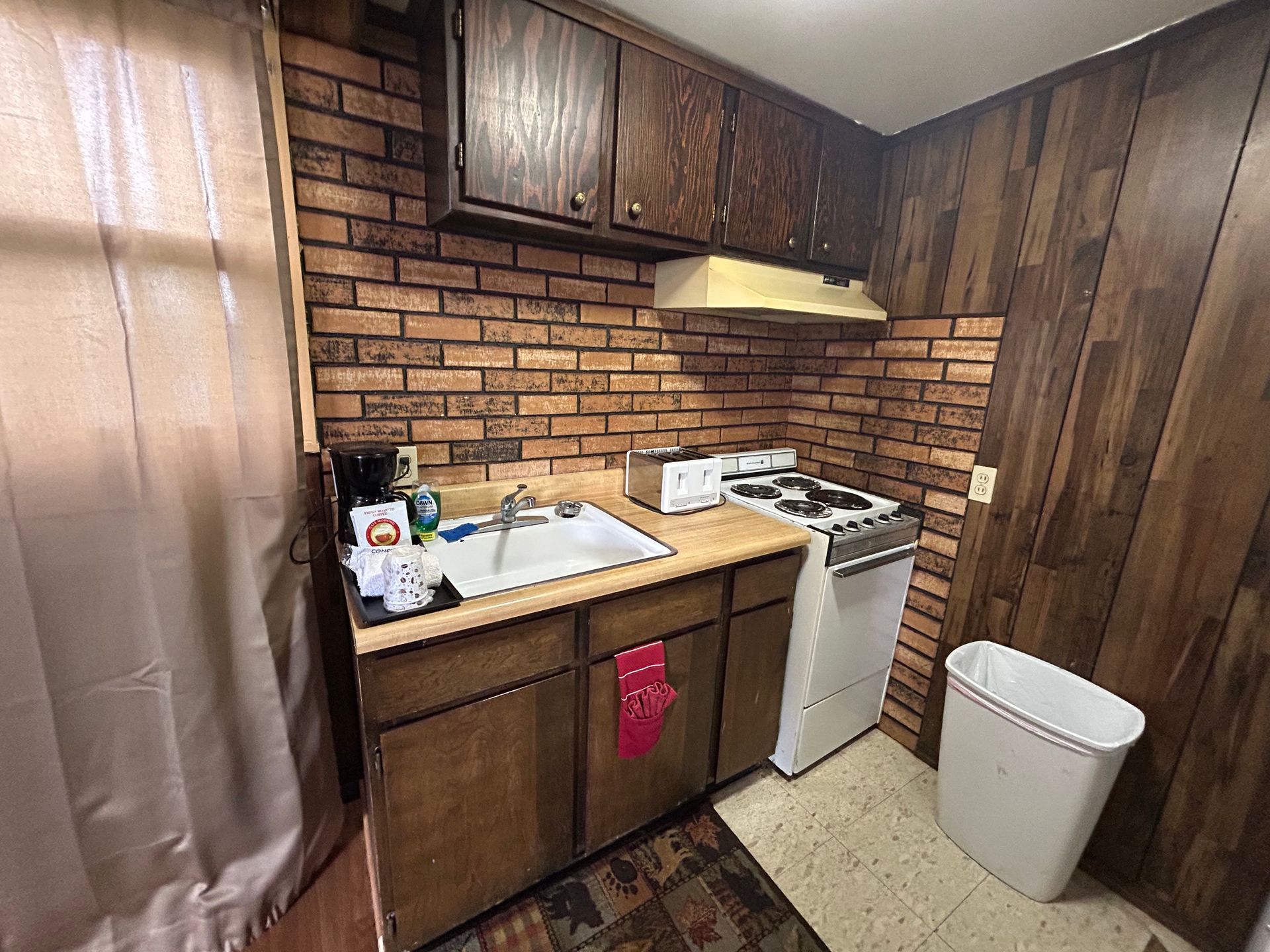 Small kitchen with brick backsplash, wooden cabinets, appliances and a trash can.