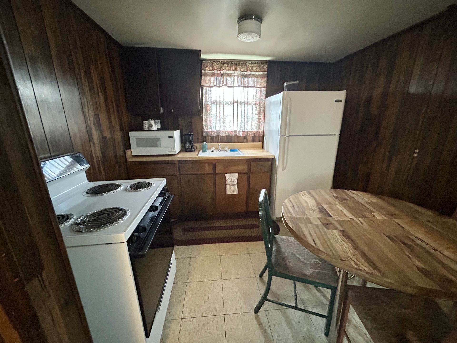 Small kitchen with wood paneling, white appliances, a round table, and a window.
