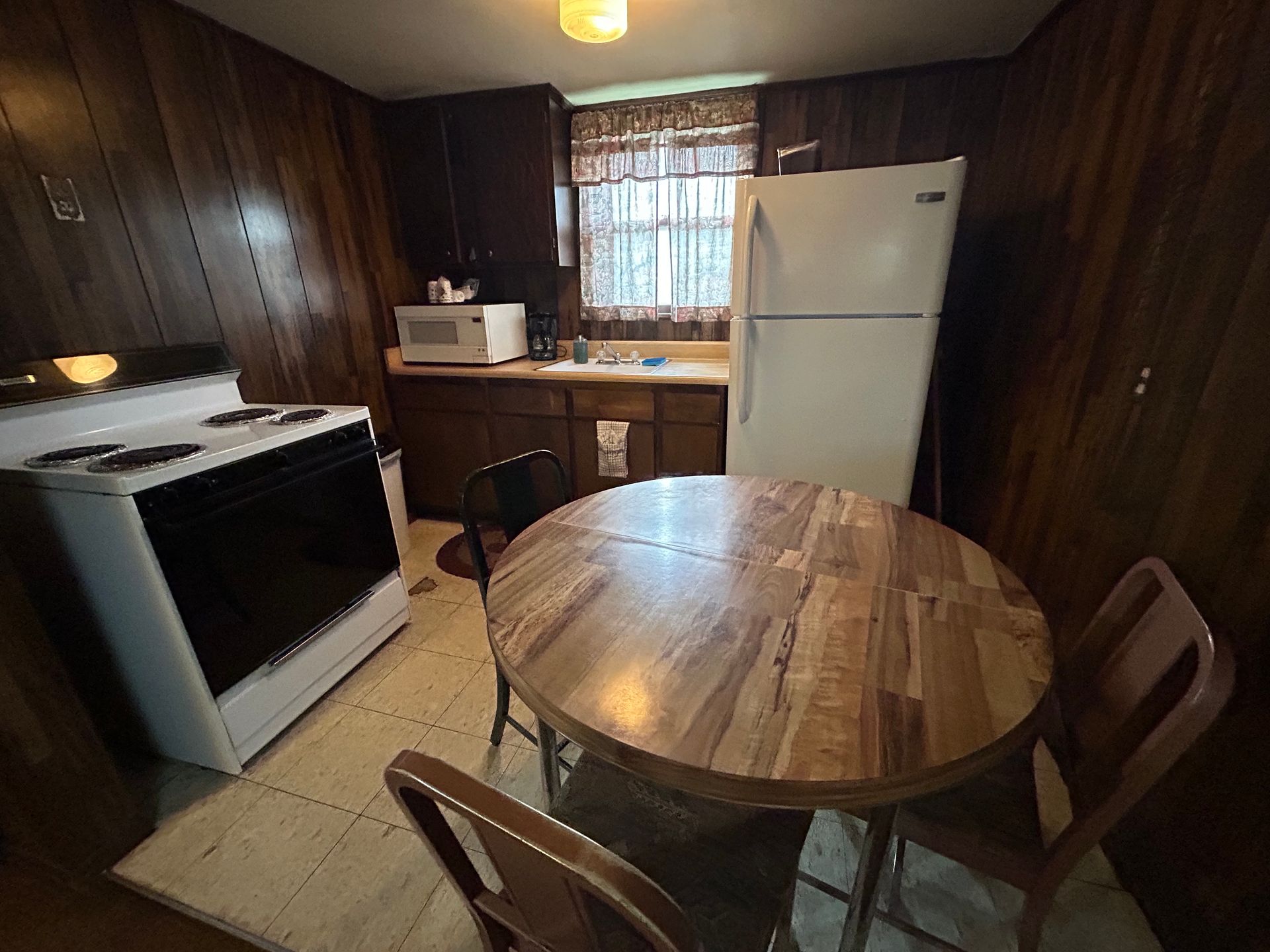Small kitchen with a stove, refrigerator, table, and dark wood paneling.