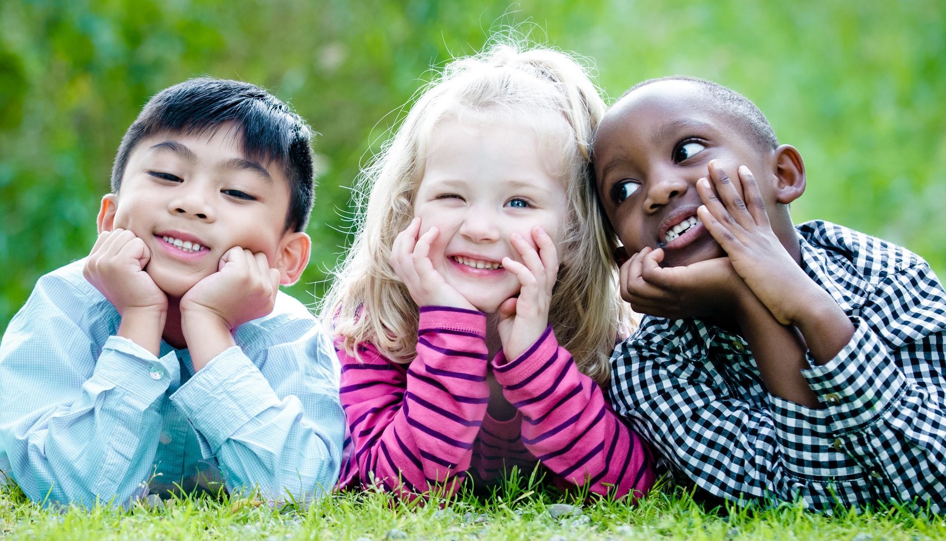 Three children lying on grass, smiling with hands on their cheeks outdoors.
